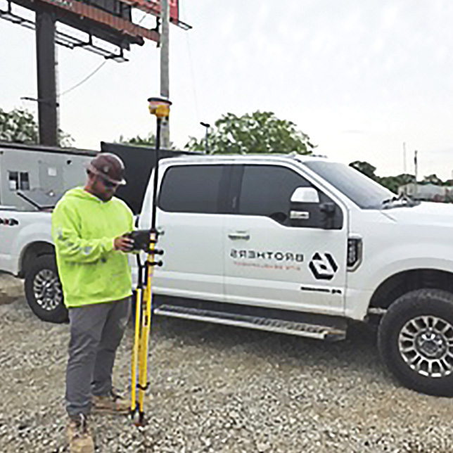 A worker in a high-visibility hoodie uses GPS land surveying equipment next to a white Brothers Site Development fleet pickup truck.
