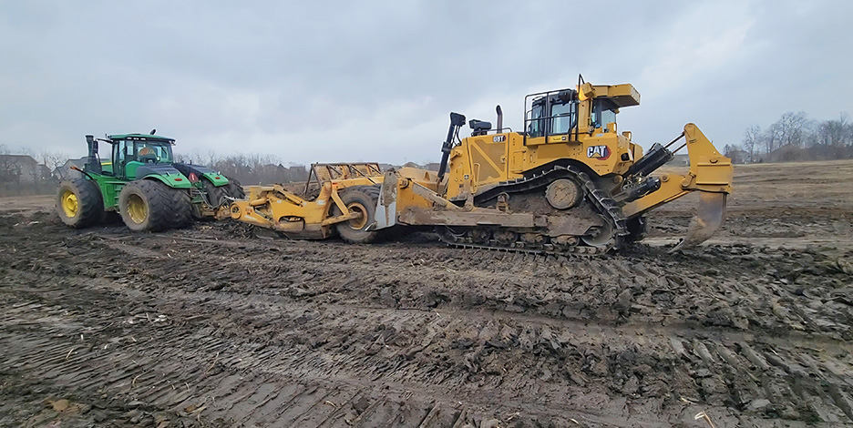 Heavy construction machinery, including a yellow bulldozer and green tractor, grading and leveling a large muddy dirt field.
