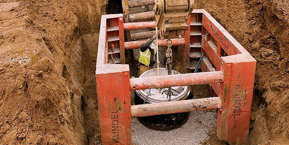 Construction worker inside a red protective trench shield installing a concrete sanitary sewer manhole in a deep dirt trench.