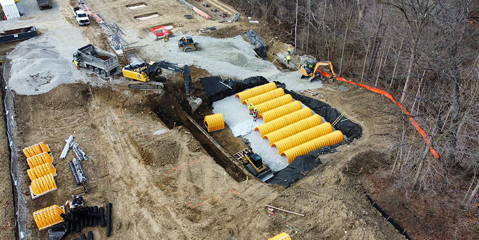 Aerial view of an excavation site where multiple large, yellow underground storm drainage retention pipes are being installed.