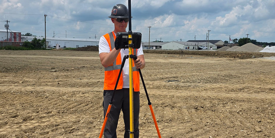 A land surveyor in a safety vest and hard hat operates GPS surveying equipment on a tripod in a wide dirt construction field.