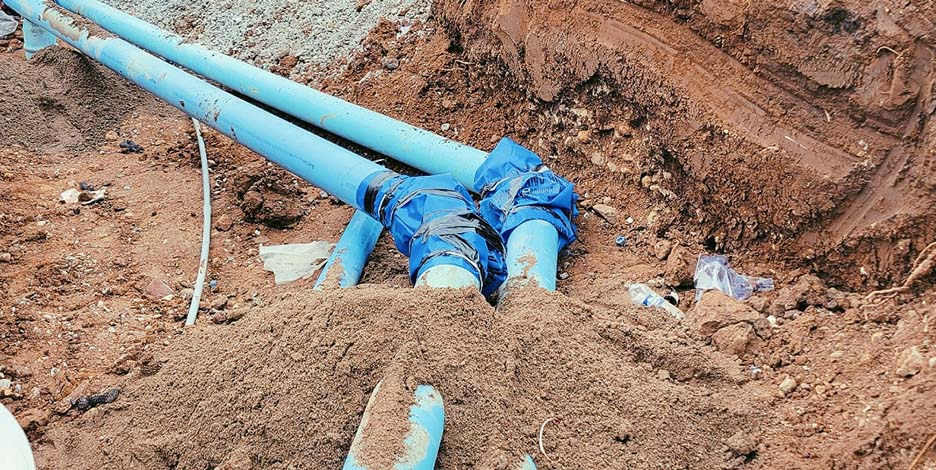 Large blue underground water main pipes and fittings laid out inside an excavated dirt trench at a construction site.